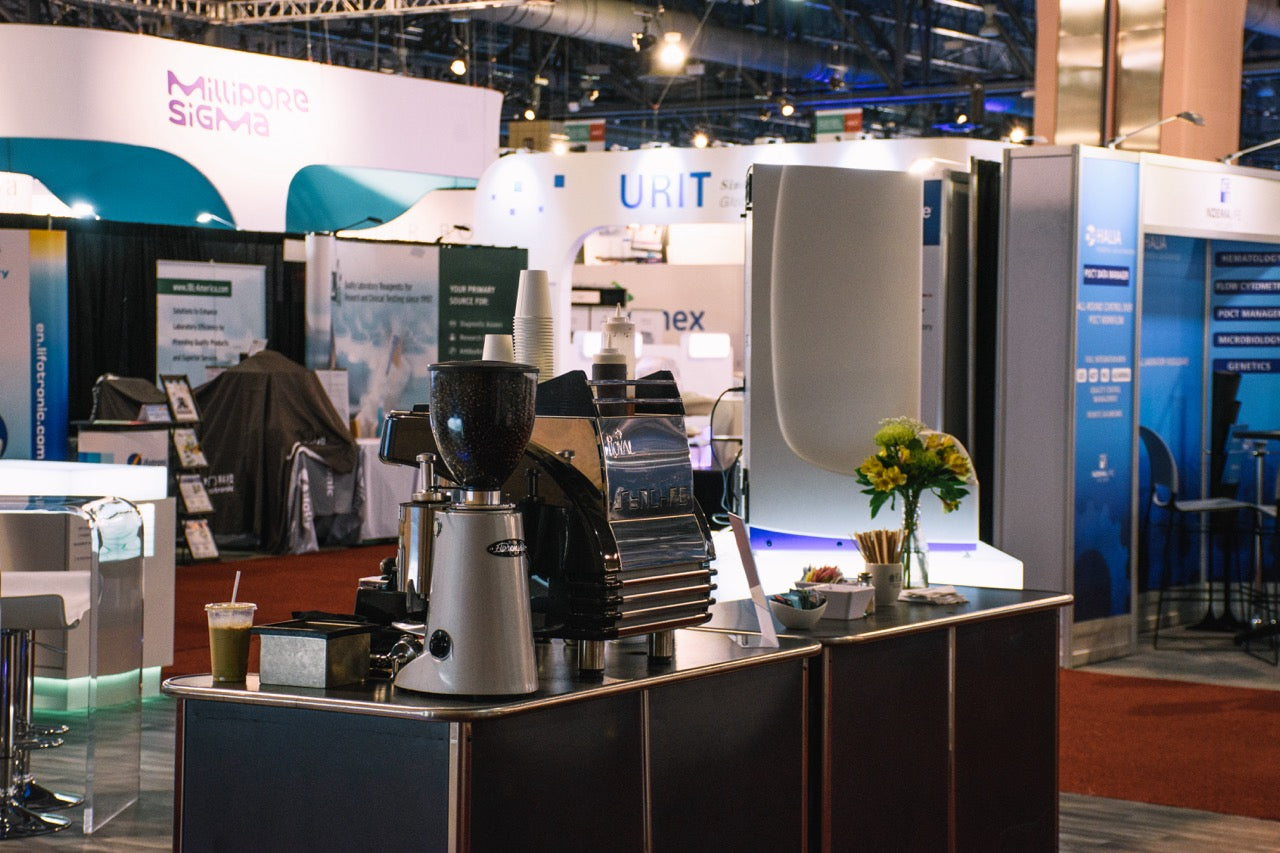 A professional espresso machine and coffee grinder set up on a catering counter at a trade show.