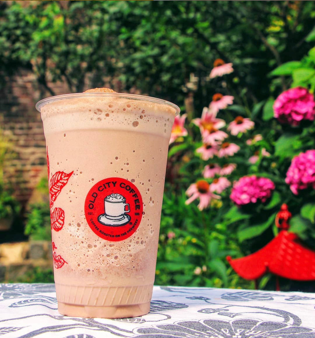 A tall, clear plastic cup filled with a frothy iced cappuccino, displaying the vintage Old City Coffee logo against a beautiful backyard garden background with blooming pink flowers.