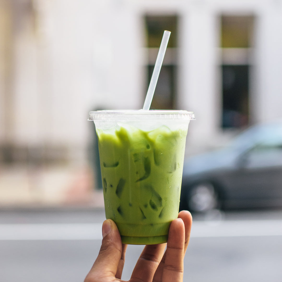 A hand holding a refreshing, bright green iced matcha green tea latte in a clear to-go cup with ice and a straw, set against a blurred urban background.