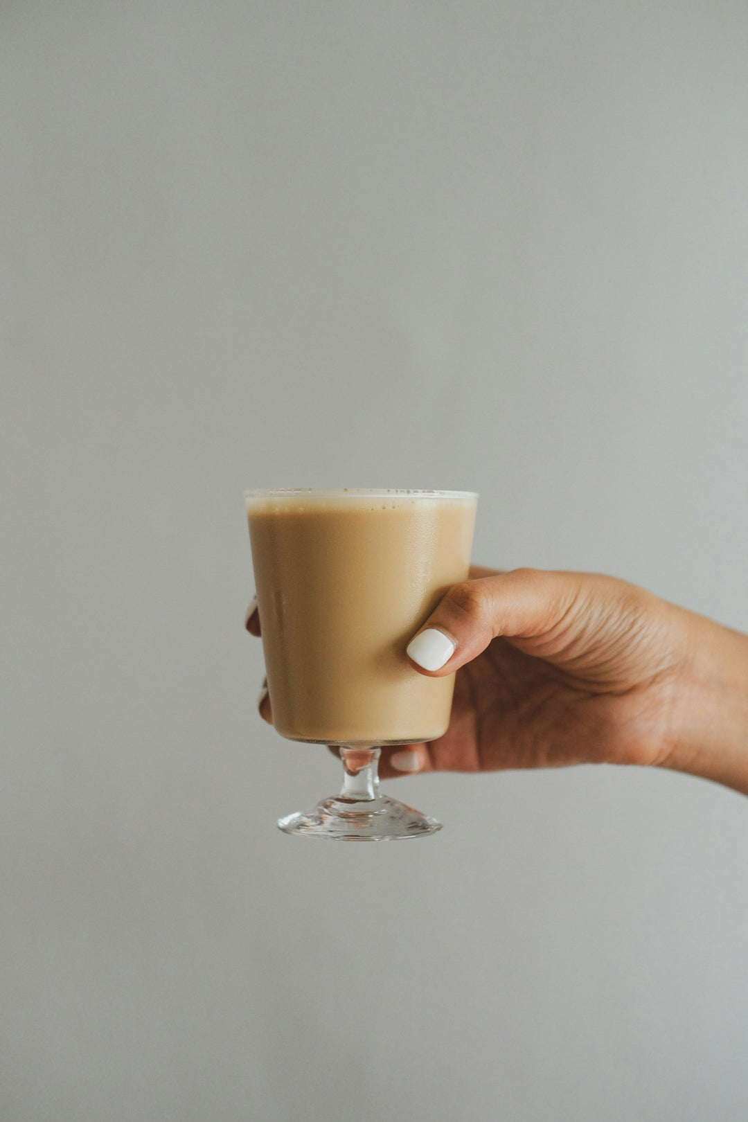A hand holding a stemmed glass of frothy, creamy nitro cold brew coffee, showcasing its rich texture against a clean, neutral background.