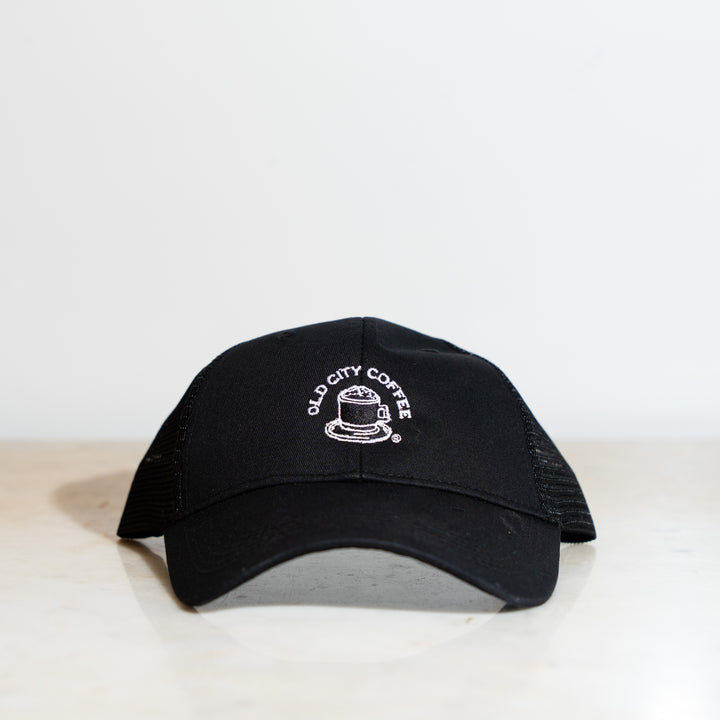 A black trucker hat with the vintage Old City Coffee logo embroidered in white. The logo features "Old City Coffee" in text that wraps around a drawing of a mug with foam.