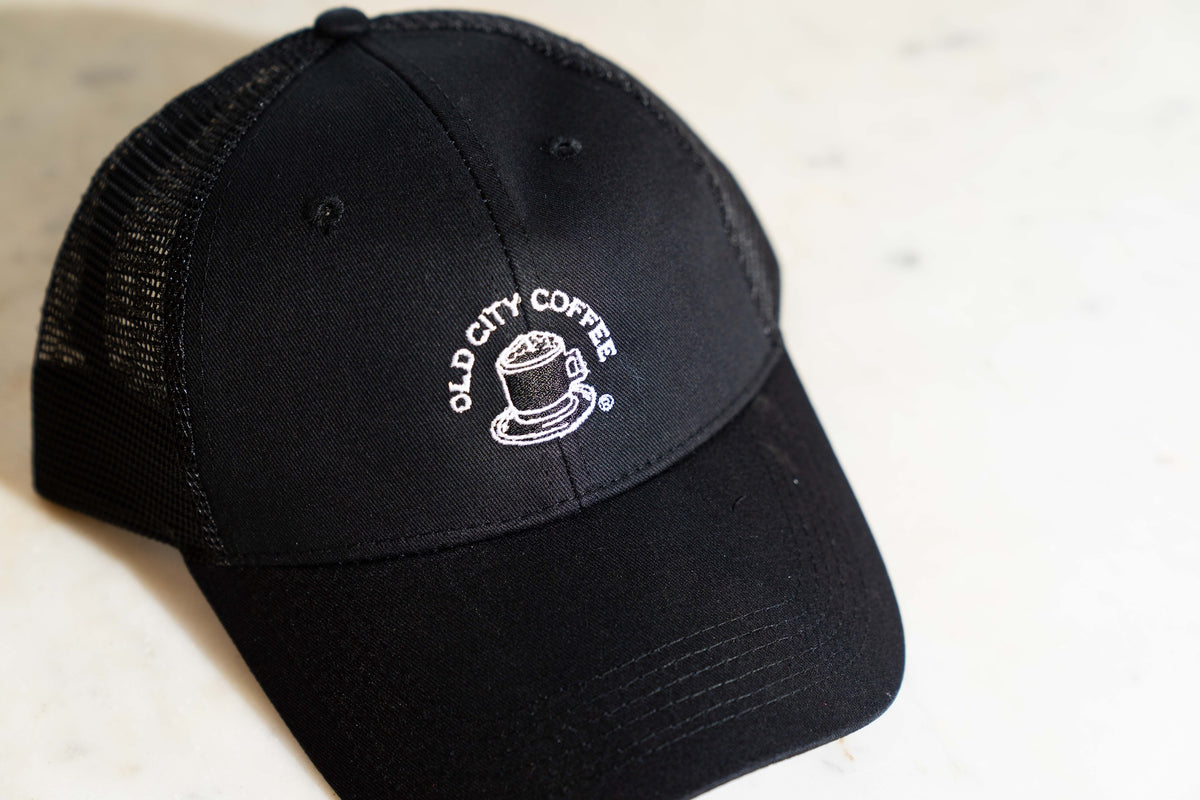 A close-up view of a black trucker hat with the vintage Old City Coffee logo embroidered in white. The logo features "Old City Coffee" in text that wraps around a drawing of a mug with foam.