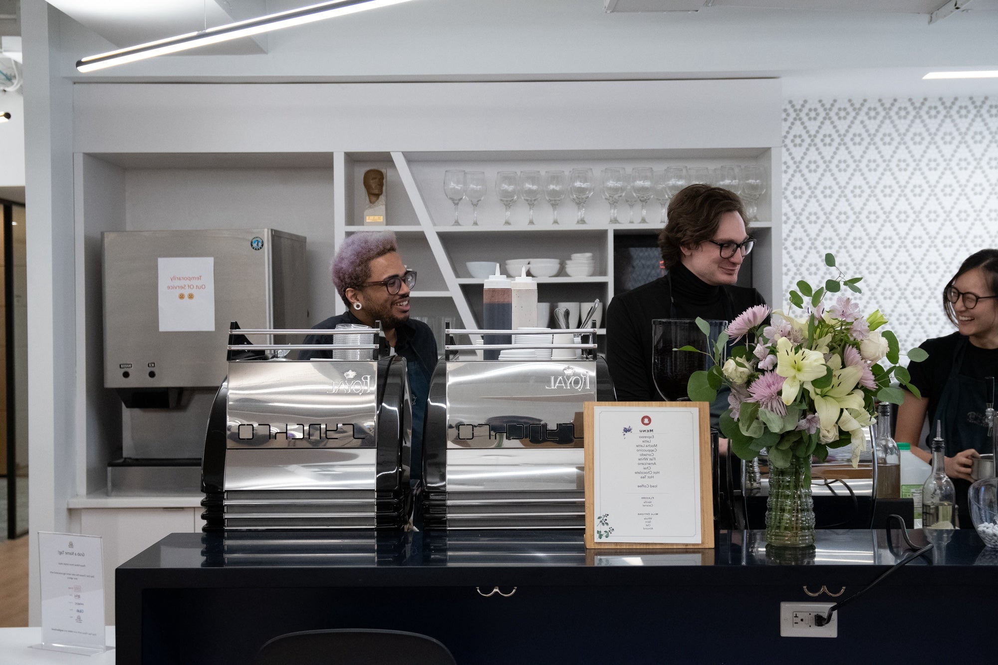 Three smiling baristas in black aprons stand behind a sleek, dark blue catering counter. On the counter are two highly polished chrome espresso machines, a wooden-framed menu, and a glass vase filled with a bouquet of white and light purple flowers. The background features white open shelving displaying glassware and a commercial coffee brewer.