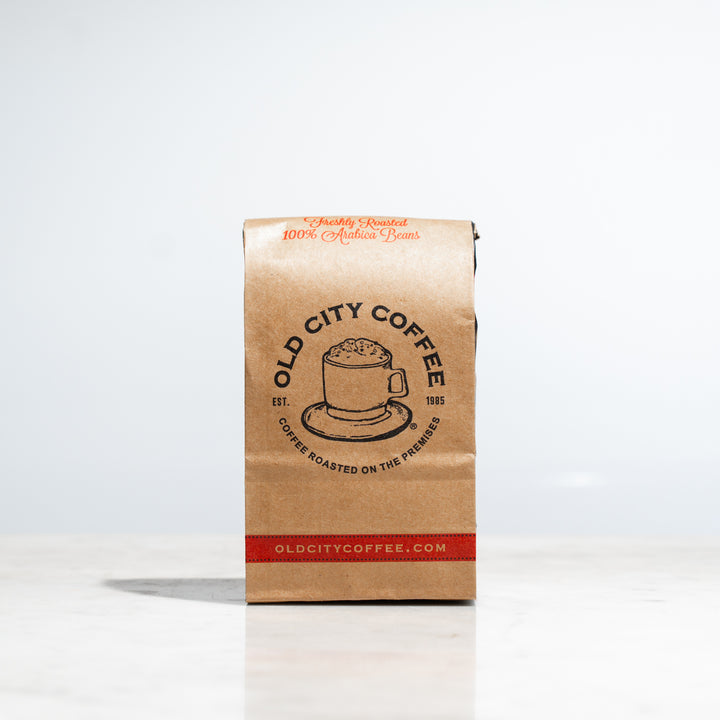 A kraft paper bag of Old City Coffee standing upright on a white marble surface against a bright white background. The bag features red text at the top reading "Freshly Roasted 100% Arabica Beans," a central circular logo with a coffee cup illustration that reads "Old City Coffee Est. 1985," and a decorative red band at the bottom with the website "oldcitycoffee.com."