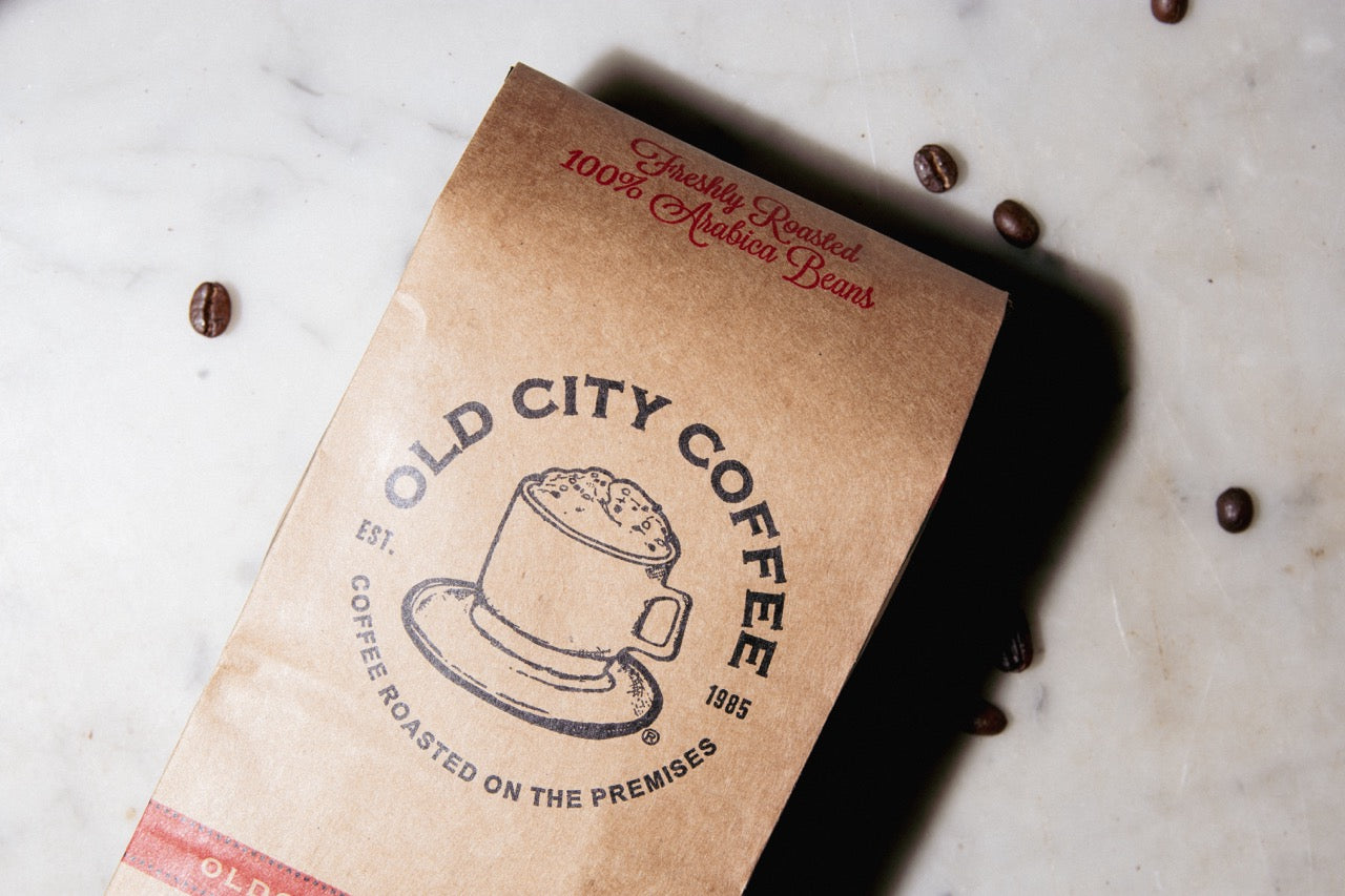 A top-down view of a brown kraft paper Old City Coffee bag lying flat on a light speckled surface. Several whole roasted coffee beans are scattered around the bag. The front of the bag displays a vintage coffee cup illustration surrounded by the text "Old City Coffee Est. 1985," and red text at the top reading "Freshly Roasted 100% Arabica Beans."