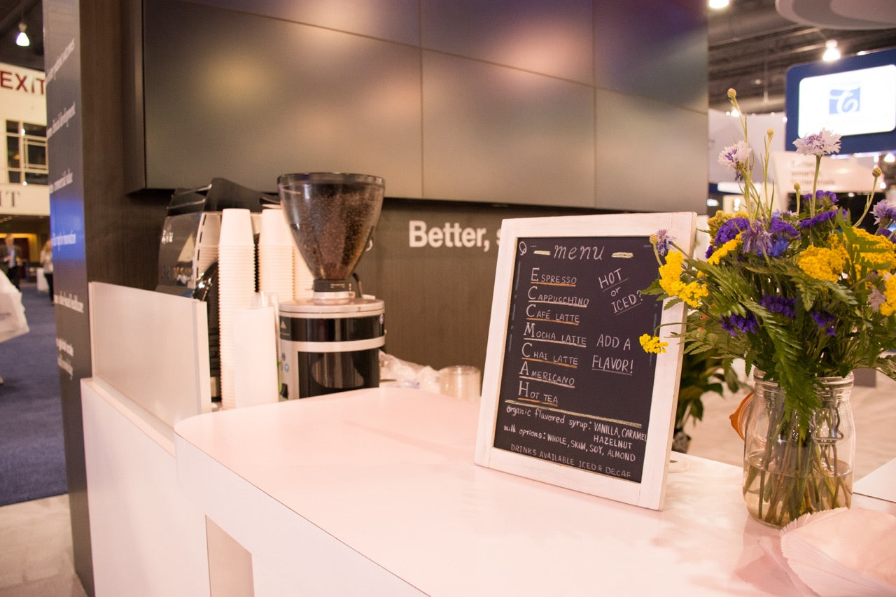 A close-up of an Old City Coffee event beverage station featuring a custom chalkboard menu, fresh flowers, and an espresso grinder on a white counter at a convention.