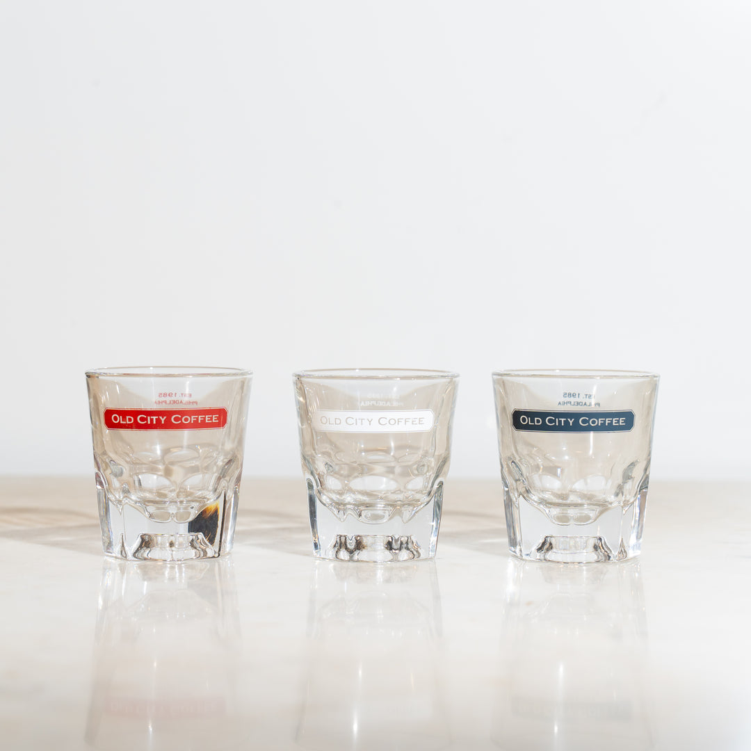 A view of three gibraltar glasses in a line featuring the Old City Coffee logo near the rim. One has the logo in red, the second has it in white, and the third has it in blue.