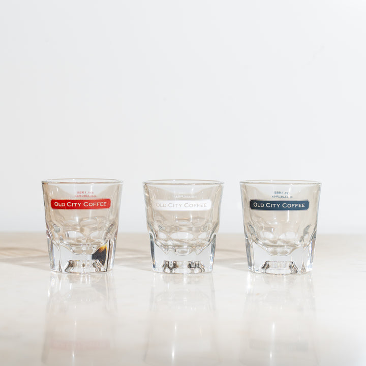A view of three gibraltar glasses in a line featuring the Old City Coffee logo near the rim. One has the logo in red, the second has it in white, and the third has it in blue.